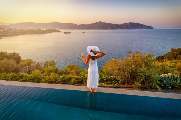 A elegant woman in a white dress stands by the swimming pool and enjoys the summer sunset behind the sea — luxury wellness Los Cabos