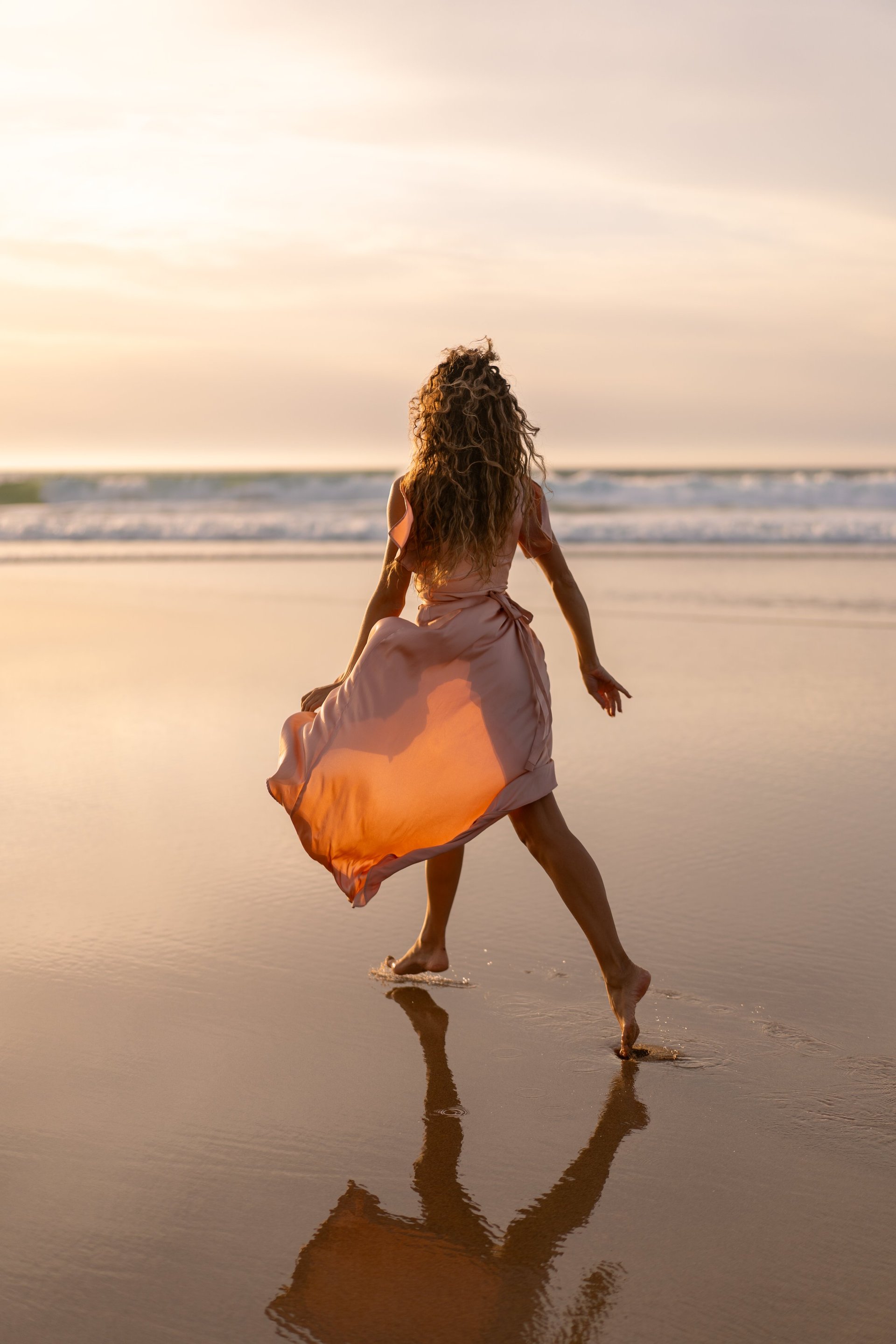 Woman in flowing dress walking barefoot on wet sand along beach at sunset