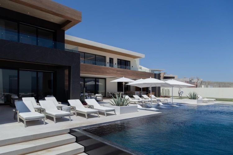 Modern luxury home with infinity pool, white lounge chairs, and contemporary architecture under clear blue sky