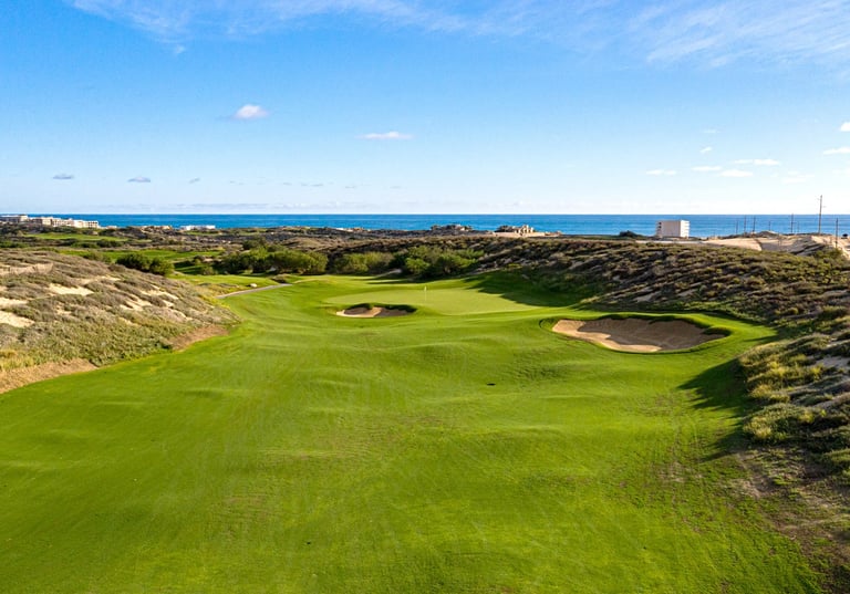 Coastal golf course hole with manicured green fairway, bunkers, and ocean view under blue sky
