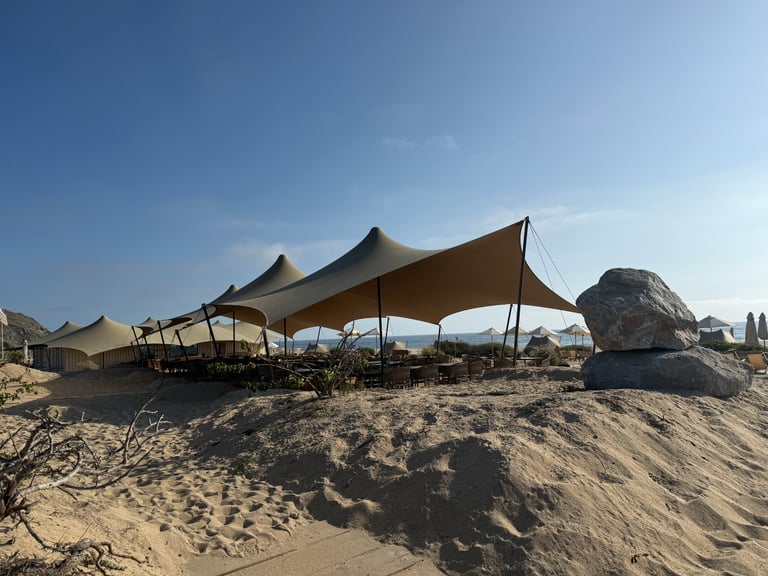 Beige tent canopies set up on sandy beach with rocks and blue sky visible in background