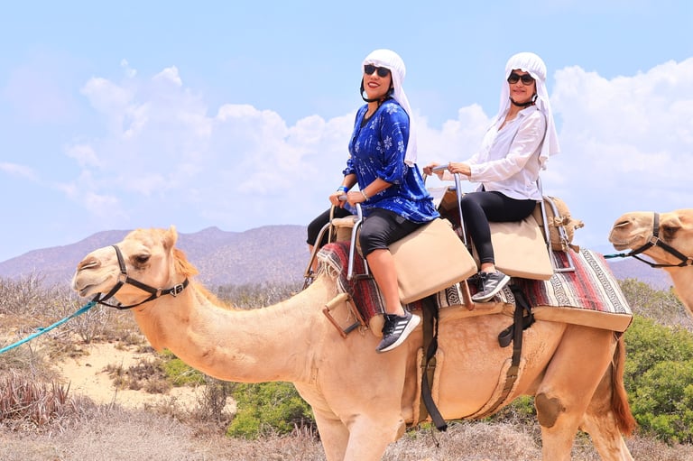 Two women wearing white hats and sunglasses ride camels in a desert landscape with mountains in the background
