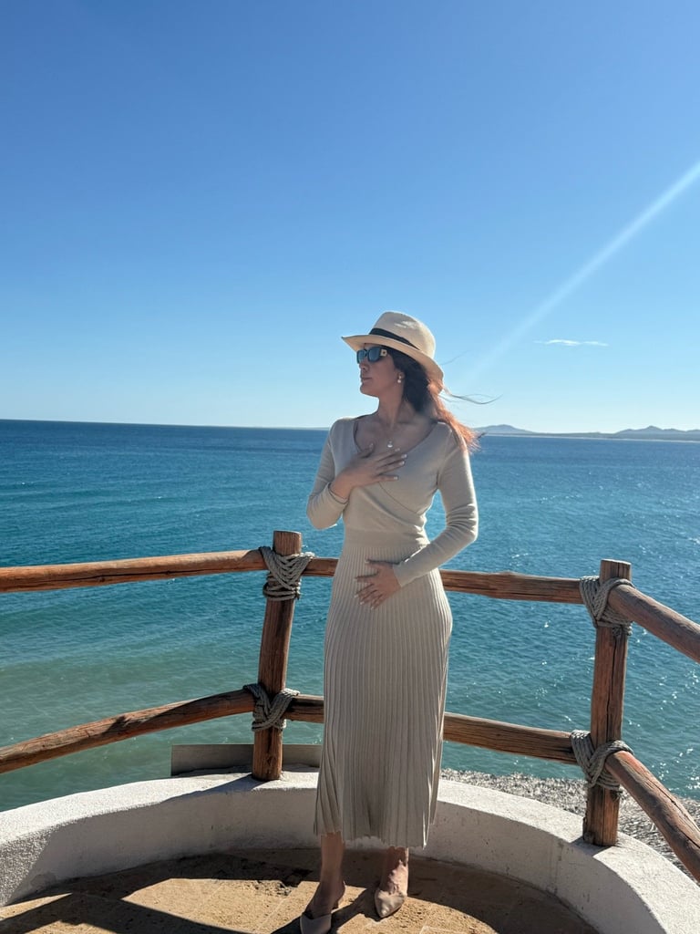 Woman in white dress and sun hat standing on wooden deck overlooking turquoise ocean water