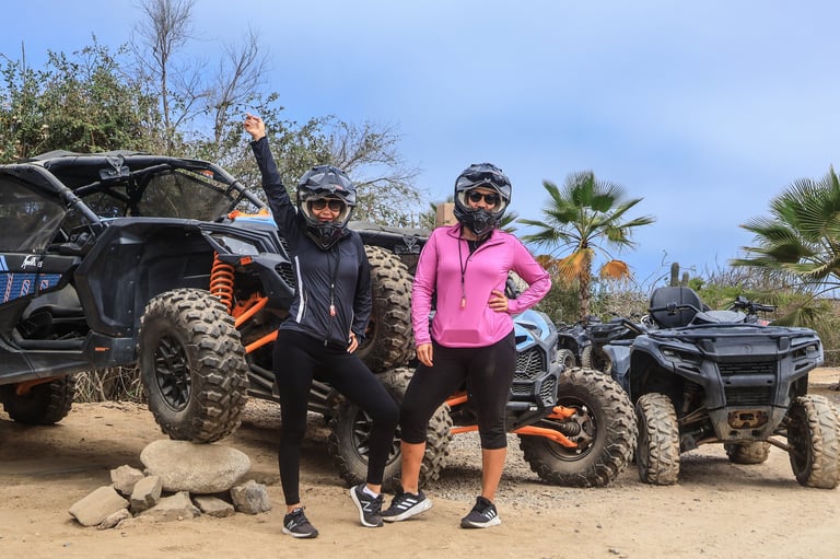 Two people in helmets posing in front of off-road vehicles in a desert setting with palm trees