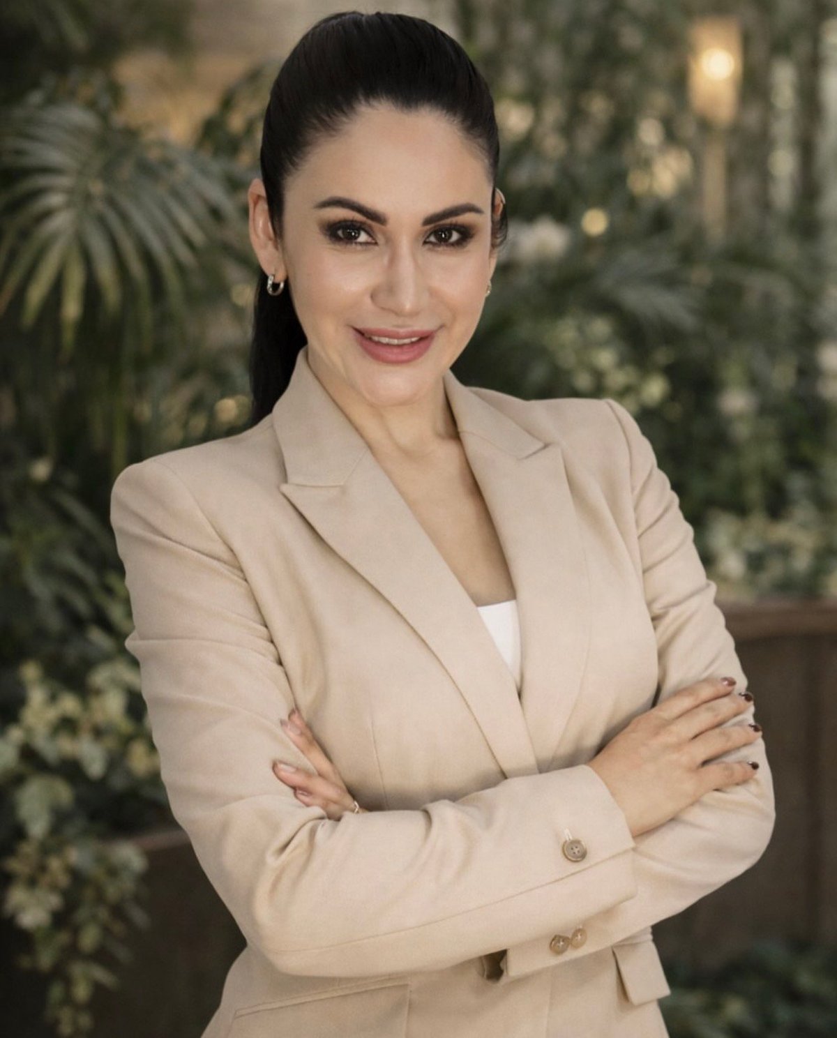 Professional woman in beige blazer with arms crossed, smiling at camera with potted plants in background