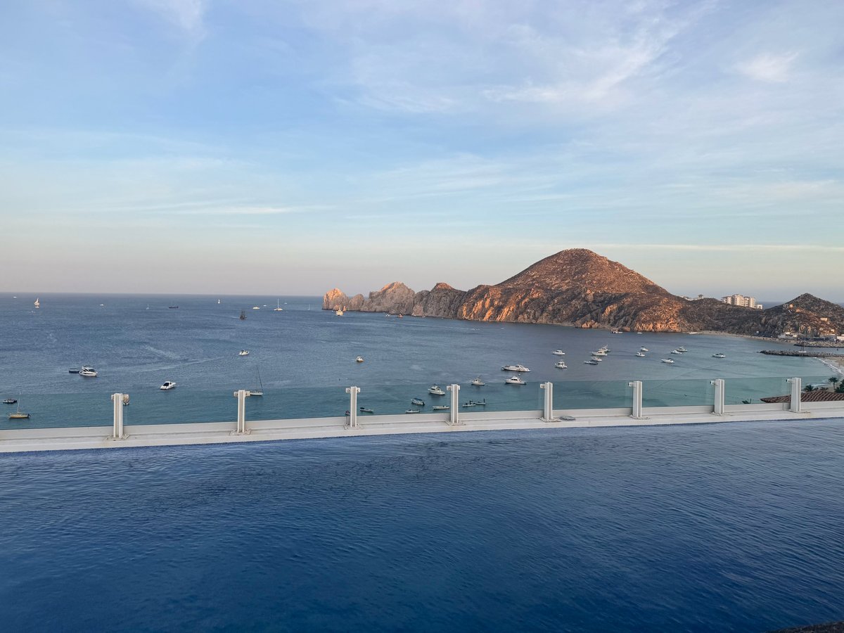 Infinity pool overlooking coastal bay with moored boats, rocky islands, and mountainous shoreline under blue sky