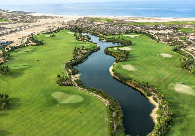 Aerial view of a scenic golf course with winding blue waterway, green fairways, and sand bunkers along a coastal landscape